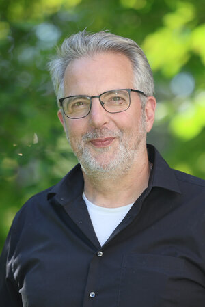 Portraitfoto Marc Leuther in Zivilkleidung vor einer grünen Buchenehecke