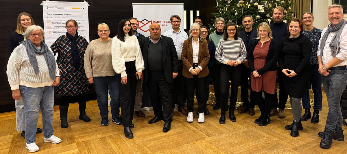 Gruppenaufnahme mit zwölf Personen in Zivilkleidung bei einem weihnachtlichen Gruppentreffen (Tannenbaum im Hintergrund)