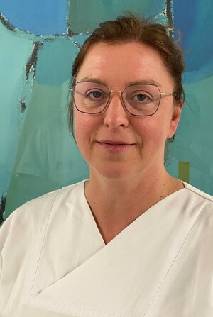 Portraitfoto einer Frau mit Brille in Dienstkleidung vor einer grün-blauen Wand