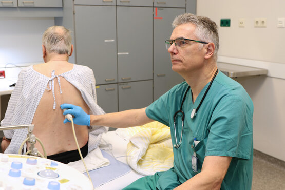 Ein Patient sitzt mit dem Rücken zu einem Arzt in grüner Dienstkleidung. Der Patient wird gerade abgehorcht