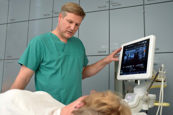 Ein Arzt in grüner Dienstkleidung bespricht mit einer liegenden Patientin, die nur von hinten zu sehen ist, einen Befund am Monitor.