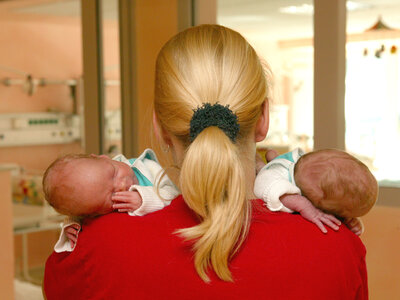 Eine blonde Pflegekraft trägt zwei kleine Säuglinge auf den Schultern und ist von hinten fotografiert